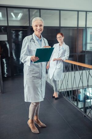 Two colleagues. Happy senior female doctor in a white coat standing in the corridor next to her smiling colleagueの写真素材