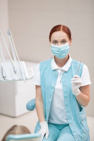 Near dental chair. Child dentist wearing mask and gloves sitting near dental chair with girlの写真素材