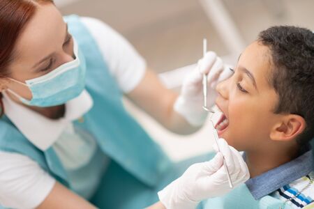 Dentist wearing mask. Red-haired child dentist wearing mask examining cute dark-skinned boyの写真素材