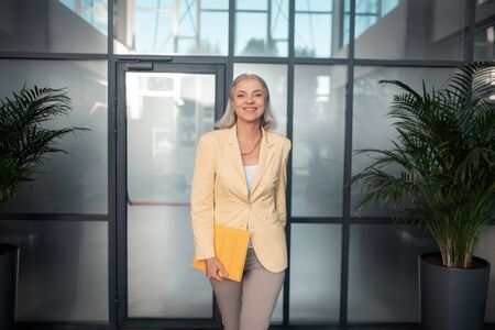 Contented look. Contented Caucasian senior businesswoman with a yellow paper folder standing in the office building and looking in front of herの写真素材
