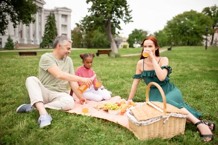 Picnic with parents. Dark-skinned daughter feeling joyful while having picnic with adoptive parentsの写真素材