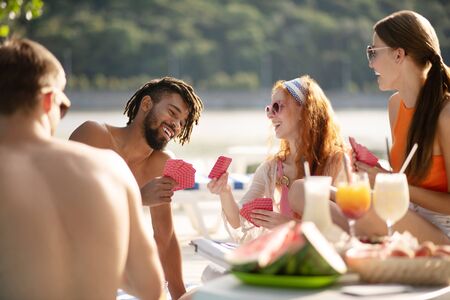 Friends playing cards. Friends laughing while playing cards sitting near river at the weekendの写真素材