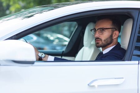 Into side mirror. Bearded man wearing hand watch looking into side mirror while driving his car