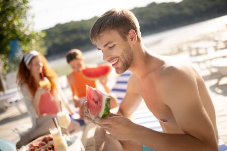 Man eating watermelon. Handsome dark-haired man smiling while eating watermelon and having restの写真素材