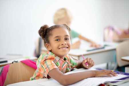 Girl feeling joyful. Dark-eyed African-American girl feeling joyful while sitting at the desk and...