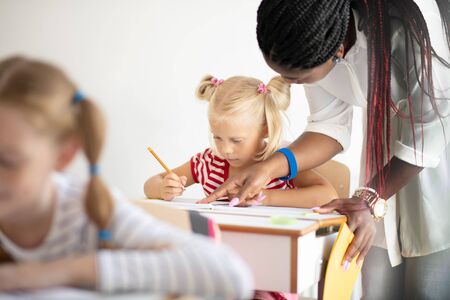 Girl listening. Blonde-haired cute little girl listening to teacher attentively and writing downの写真素材