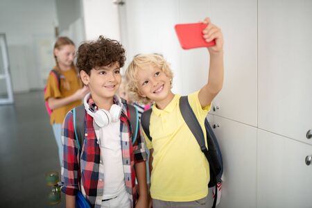 Selfie with classmate. Handsome cheerful boy holding red smartphone while making selfie with classmateの写真素材