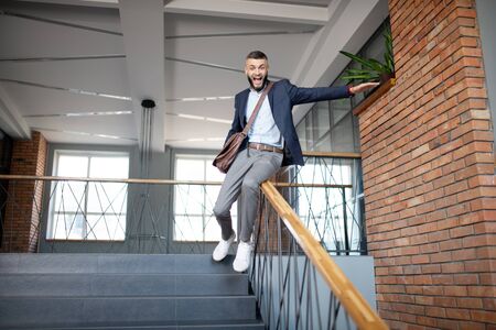 Sliding on banister. Cheerful bearded dark-haired man having fun while sliding on the banisterの写真素材