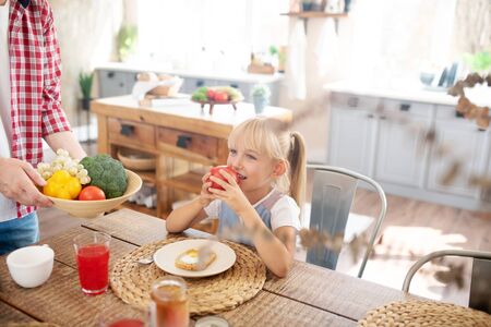 Daughter biting tomato. Cute blonde daughter biting tomato while having breakfast early in the morningの写真素材