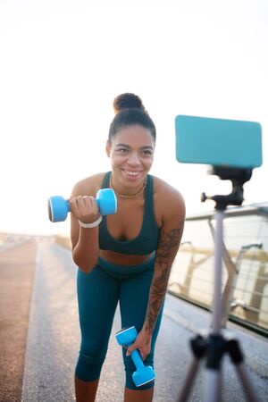Exercises with barbells. Dark-skinned woman doing exercises with barbells while filming blogの写真素材