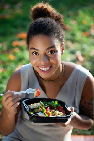 Yummy vegetable salad. Dark-eyed woman smiling while eating yummy vegetable salad sitting on grassの写真素材