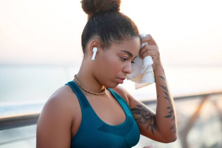 Woman feeling exhausted. Dark-skinned young woman taking towel while feeling exhausted after long runの写真素材