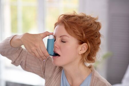 Having asthma attack. Young red-haired woman using inhalator while having asthma attack