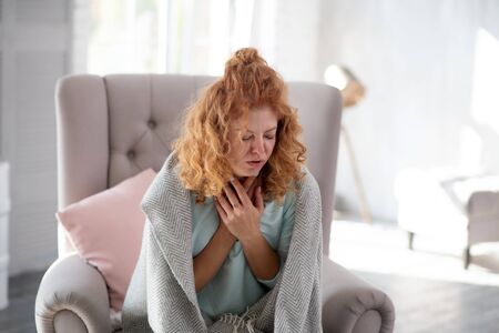 Woman coughing. Curly red-haired woman coughing while sitting on armchair having sick leaveの写真素材