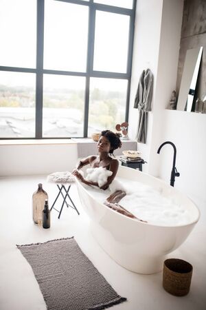 Having foamy bath. Naked dark-skinned woman feeling good while having foamy bath at the weekendの写真素材