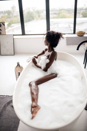 Looking into window. Curly dark-skinned woman looking into window while lying in bath with foamの写真素材