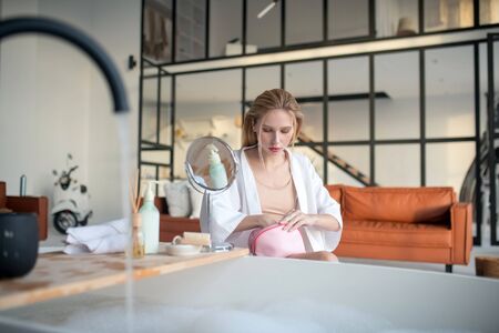 Sitting at table. Blonde woman wearing white bathrobe sitting at the table and doing makeup
