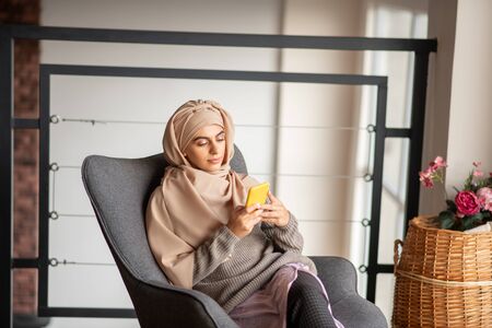 Spacious cozy room. Pretty woman with dark eyes using phone while sitting in the spacious cozy room