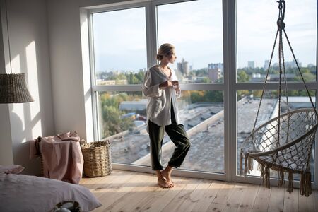 In living room. Blonde woman wearing baggy trousers standing near window in living roomの写真素材