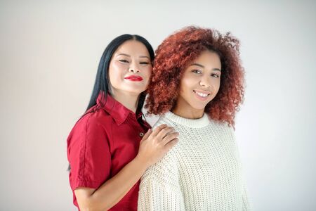 Standing near friend. Curly red-haired woman standing and hugging near her Asian excited friendの写真素材