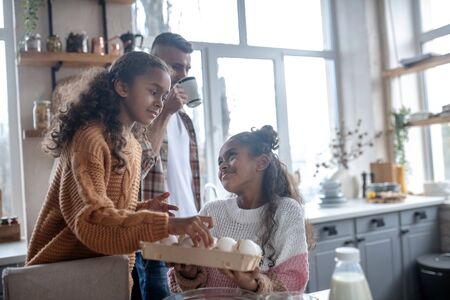 Sister giving eggs. Cute sister giving eggs while cooking traditional family pie with fatherの写真素材