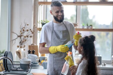 Father taking detergents. Beaming father taking detergents while cleaning kitchen with daughterの写真素材