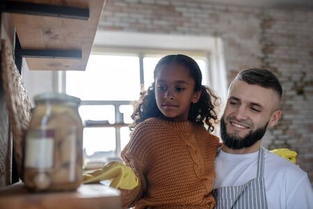 Daughter dusting shelves. Bearded father smiling while daughter dusting shelves in the kitchen