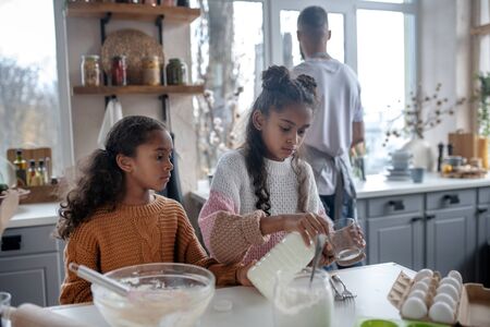 Cooking with sister. Appealing younger sister pouring milk into glass while cooking pie with sisterの写真素材