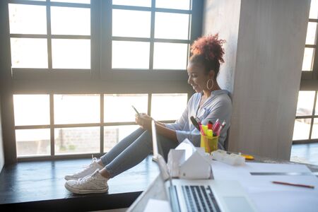Sitting near window. Interior designer having break while sitting near window and using phone