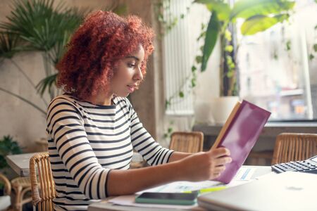 Business literature. Red-haired young businesswoman reading business literature while sitting in...