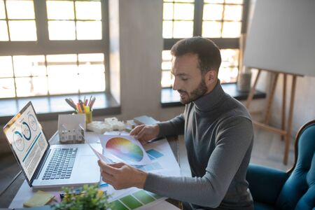 Sitting near laptop. Handsome young interior designer sitting near laptop and looking at color paletteの写真素材