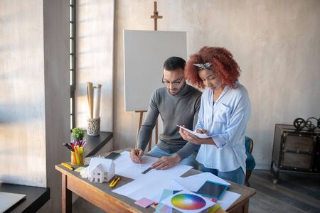 Near interior designer. Curly assistant making notes while standing near interior designer making...