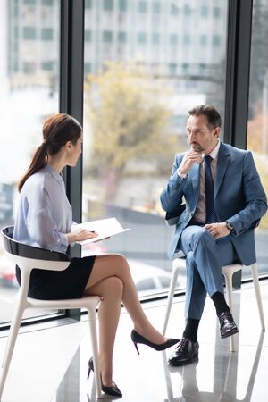 Enjoying talk. Rich entrepreneur enjoying talk with young experienced journalist while sitting near the windowの写真素材
