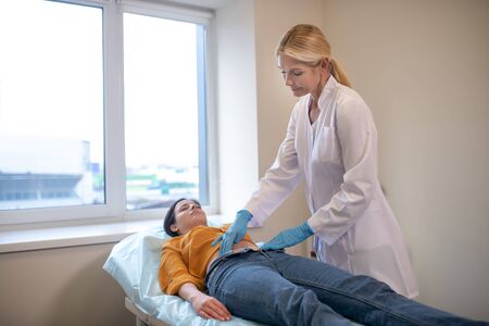 Stomach test. Blond doctor looking serious wearing sterile gloves palping patients stomach