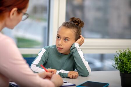 Being unhappy. Little schoolgirl sitting leaning on her arm and looking at her teacherの写真素材
