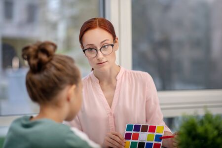 Colors and mood. Serious teacher showing a schoolgirl a sheet of paper with colourful tableの写真素材