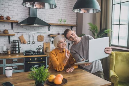Meeting online. Young handsome man in grey shirt and his mom talking online with friendsの写真素材