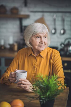 In the kitchen. Good-looking elderly lady in yellow sitting in her kitchenの写真素材