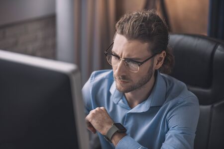 Looking serious. Young bearded man in blue shirt working at the computerの写真素材