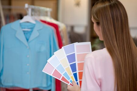 Work with color scales. Young designer with color scales in her hands, located in the showroom opposite the coat rack with a collection of clothes, looking at the clothes.の写真素材