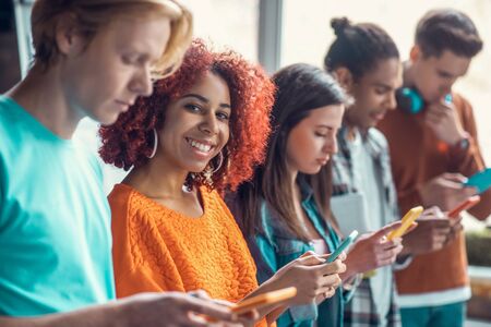 Using social media. Curly red-haired student smiling standing near friends and using social media