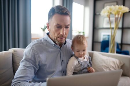 Busy father. Mature father holding his kid and working on his laptop
