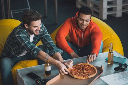 So hungry. Handsome hungry men sitting together on the sofa while taking pieces of pizzaの写真素材