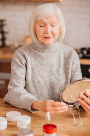 Mirror. Grey-haired beautiful lady in grey blouse holding a mirrorの写真素材