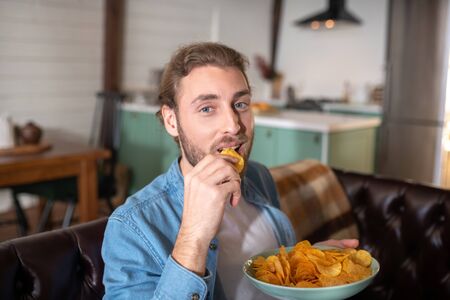 Eating habits. A joyful amn eating tasty chips at homeの写真素材