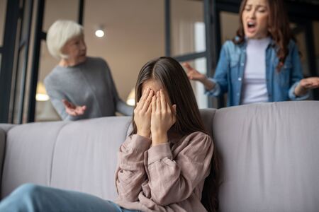 Difficult age. Unhappy daughter covering her face with hands sitting on the couch, mother and grandmother standing behind worried talking arguments.の写真素材