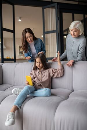 Parental control. Serious little girl sitting with a smartphone in her hand and looking attentively at the screen, grandmother with mom are distressed standing behind.の写真素材