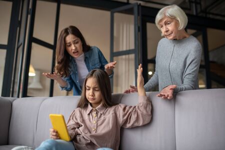 Difficult relationship. Daughter, carried away by a smartphone, looking at the screen, stopping the conversation of her grandmother and mother, who are upset.の写真素材