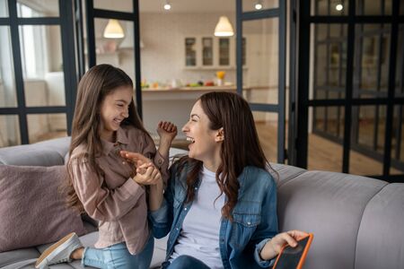 Funny moment. Beautiful and happy mother and daughter sitting on the couch, laughing, looking at each other, in a great mood.の写真素材
