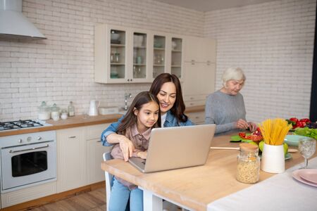 In the kitchen. Young pretty woman with a joyful daughter working at a laptop sitting at a table,...